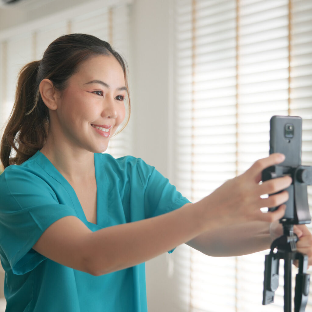 A nurse in scrubs smiling while video calling on a smartphone.
